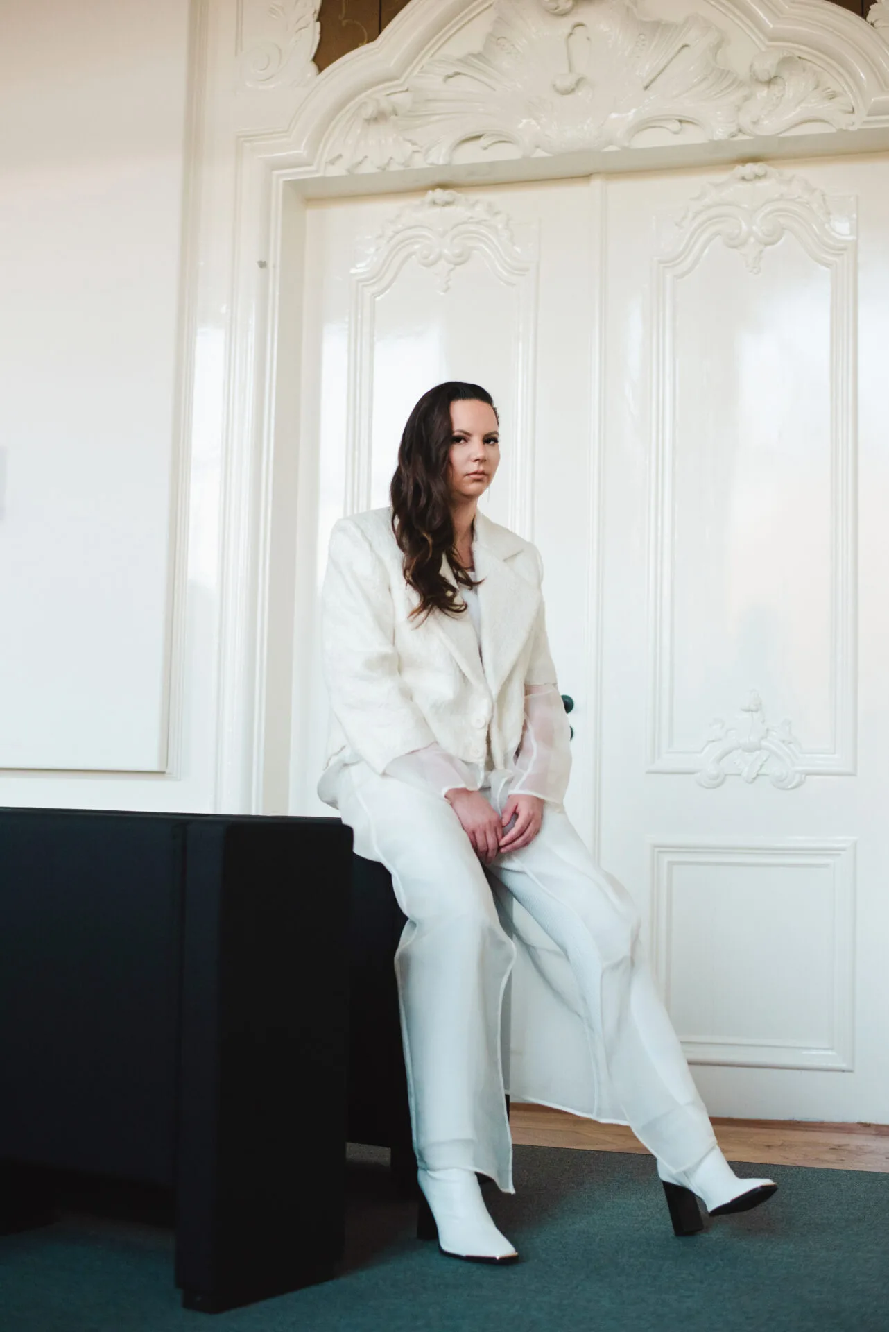Founder Daisy sitting half on a table wearing a white suite with white heels, with hair on 1 side facing the camera